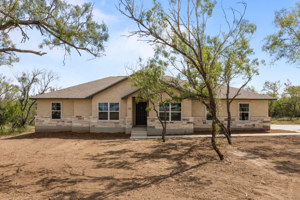 A tan single-story house with light stone accents, surrounded by slender trees and an open dirt yard, set under a bright blue sky.