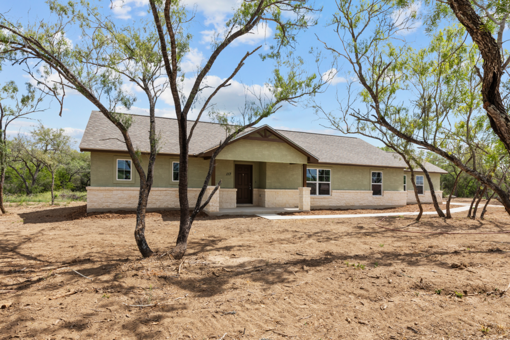 A single-story house with a green exterior and stone accents, surrounded by sparse trees and open land under a partly cloudy sky.